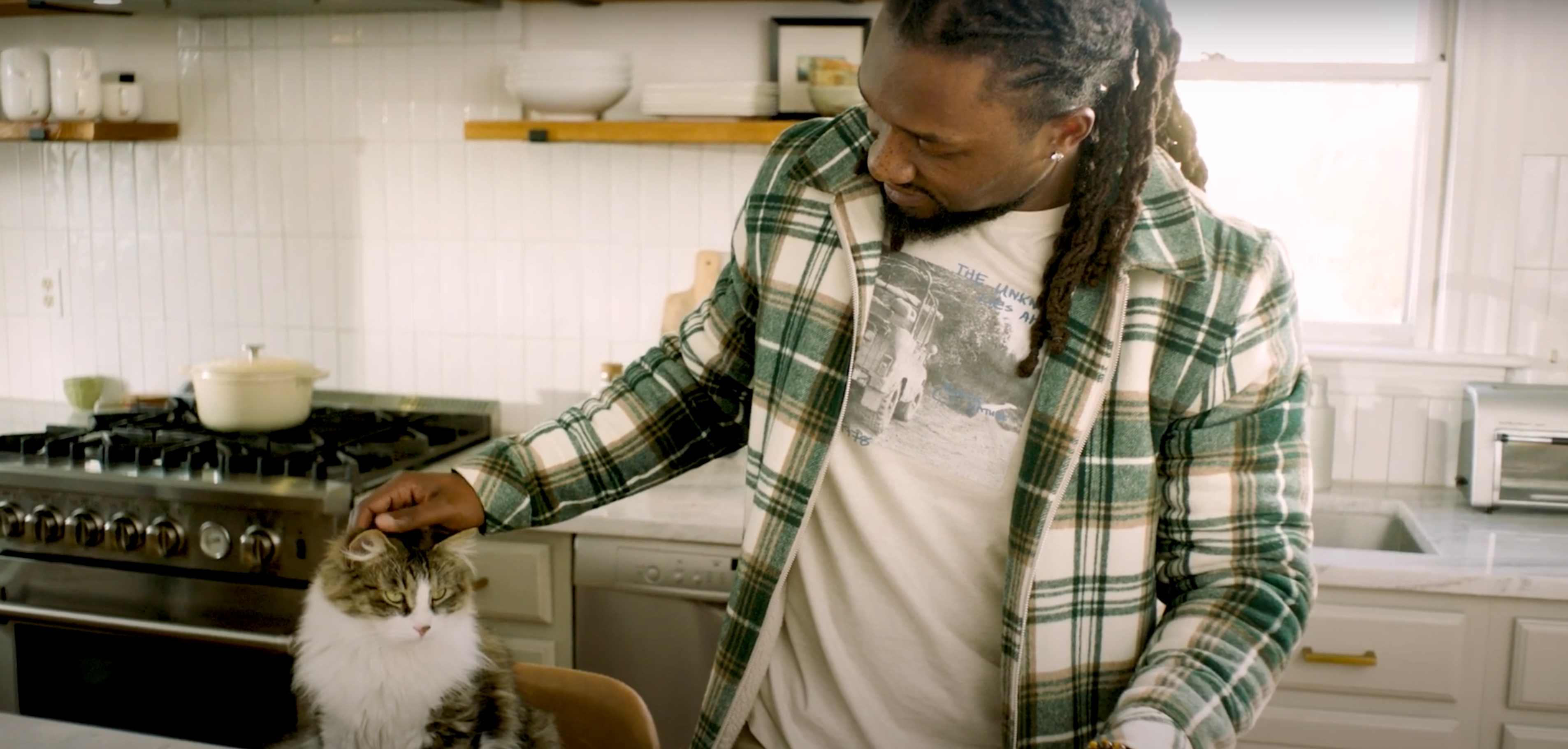 Man in kitchen petting his cat sitting on a stool.  Cat looks relaxed as he gets his head patted.